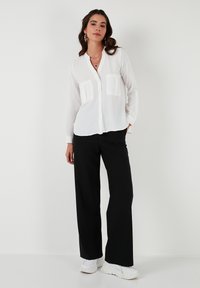 White long-sleeve shirt with button front and two patch pockets, paired with loose-fitting black pants and white sneakers. Minimalist design.