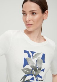 Witte katoenen t-shirt met een blauw en zilver grafisch ontwerp van bladeren. Opstaande kraag en korte mouwen met een gladde textuur.
