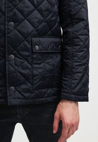 Quiltad mörkblå jacka i syntetmaterial med knappstängning, två framfickor och strukturerat diamantmönster.