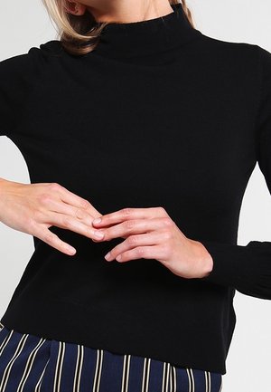 Kobieta ubrana w czarną bluzkę z długim rękawem i paskowane spodnie, z rękami delikatnie stykającymi się na wysokości talii.