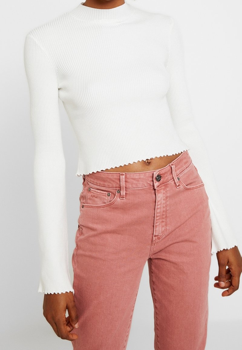 Weißes, geripptes Crop-Top mit langen Ärmeln und wellenförmigem Saum, kombiniert mit pinken, hoch taillierten Jeans. Glatte Textur und figurbetontes Design.