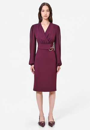 Femme portant une robe prune longueur genou avec des manches longues transparentes, une large ceinture avec un détail en chaîne dorée et des chaussures pointues assorties.