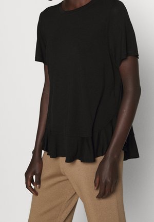 Personne portant un haut noir à manches courtes avec un ourlet à volants et un pantalon beige côtelé sur un fond clair uni.