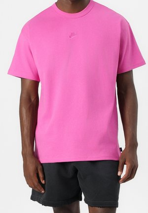 Mann trägt ein knallpinkes Nike-T-Shirt und schwarze Shorts, steht vor einem einfachen weißen Hintergrund.