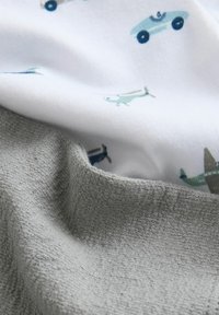 Tissu gris doux recouvrant partiellement un tissu blanc avec un motif de petits avions et voitures jouets bleus et verts.