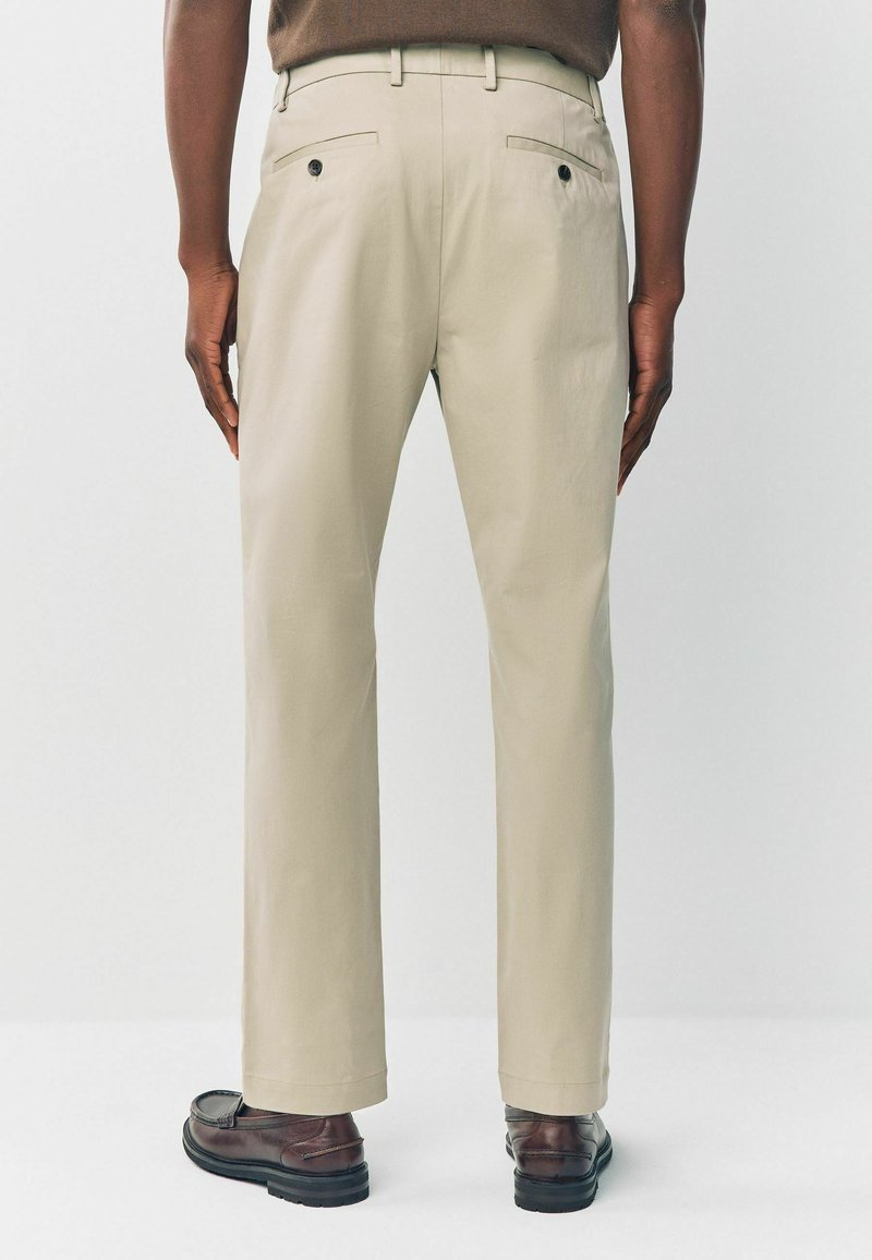 Beige Hosen, von hinten dargestellt, mit einer glatten Textur, zwei Gesäßtaschen mit Knöpfen und gesäumten Säumen, kombiniert mit braunen Schuhen.