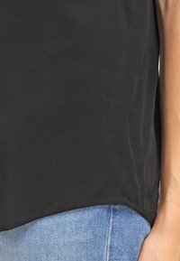 Top in tessuto nero e liscio con orlo arrotondato. Il bordo inferiore è leggermente più lungo sul retro. Indossato sopra jeans in denim azzurro chiaro.