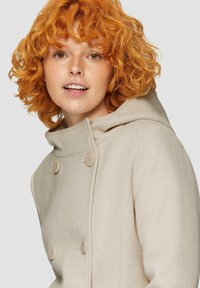 Lichtbeige dubbelzijdige jas met een capuchon, voorzien van grote neutrale knopen en een zachte, gestructureerde stof. Krullend oranje haar zichtbaar.