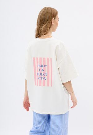 Oversized biały T-shirt z różowo-białym wzorem w paski na plecach, z niebieskim napisem "ENJOY LA DOLCE VITA." Krótkie rękawy.