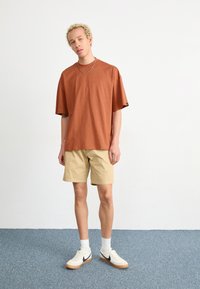 Hrdzavé oversized tričko s krátkym rukávom v kombinácii s béžovými šortkami. Oblečenie má voľný strih a je doplnené o biele tenisky a členkové ponožky.