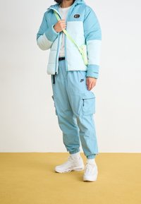 Ljusblå pufferjacka med vita detaljer och en svart logotyp, tillsammans med matchande cargo-byxor. Vita sneakers och en grön axelväska.