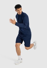 Marineblauwe sportjack met hoge kraag, gecombineerd met marineblauwe shorts. Draag het met witte sneakers en crew-sokken, op een lichtgrijze achtergrond.