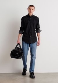 Svart skjorte med knapper og prikkete mansjetter, blå jeans, svarte sko og en svart duffelbag med et sirkulært aksent. Enkel bakgrunn.