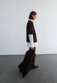 Brun oversized top lagd over en hvid skjorte, parret med brune bukser. Stoffet ser tekstureret ud, og har en slank silhuet og minimalt design.