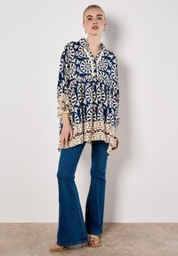 Tunik berwarna biru navy dengan pola floral putih, dilengkapi dengan kerah berbutton dan lengan berbulu, dipadukan dengan jeans biru yang melebar.
