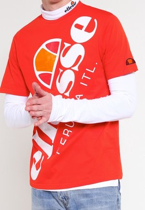 Osoba nosząca jaskrawo pomarańczowy t-shirt z logo Ellesse na białej koszuli z długim rękawem i jasnoniebieskich dżinsach, ręce splecione z przodu.