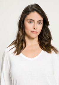 Bílá dlouhá rukávová košile s výstřihem do V a hladkým povrchem. Model má vlnité, tmavě hnědé vlasy. Pozadí je jednoduché v světlé barvě.