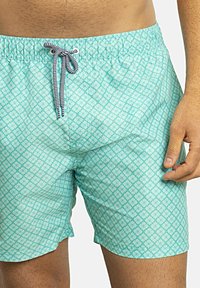 Bañadores de color turquesa claro con un patrón geométrico en blanco, cinturilla elástica y cordón negro y blanco. Textura de tela suave.