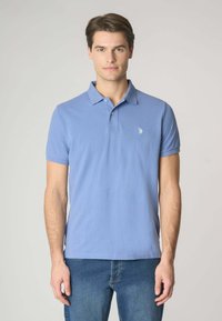 Polo in cotone azzurro chiaro a maniche corte, con colletto a tre bottoni e un piccolo emblema sul lato sinistro del petto. Indossato con jeans blu.