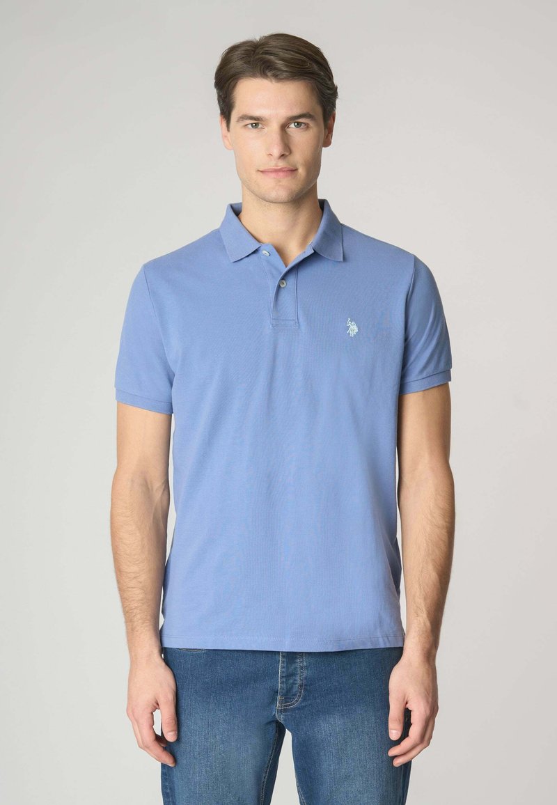 Polo in cotone azzurro chiaro a maniche corte, con colletto a tre bottoni e un piccolo emblema sul lato sinistro del petto. Indossato con jeans blu.