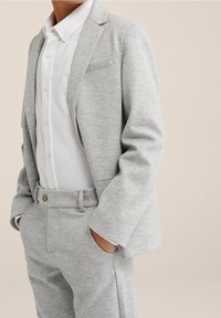 Person in einem hellgrauen Sakkowestens und einer hellgrauen Hose über einem weißen Hemd mit Knöpfen, die mit den Händen in den Taschen vor einem neutralen Hintergrund steht.