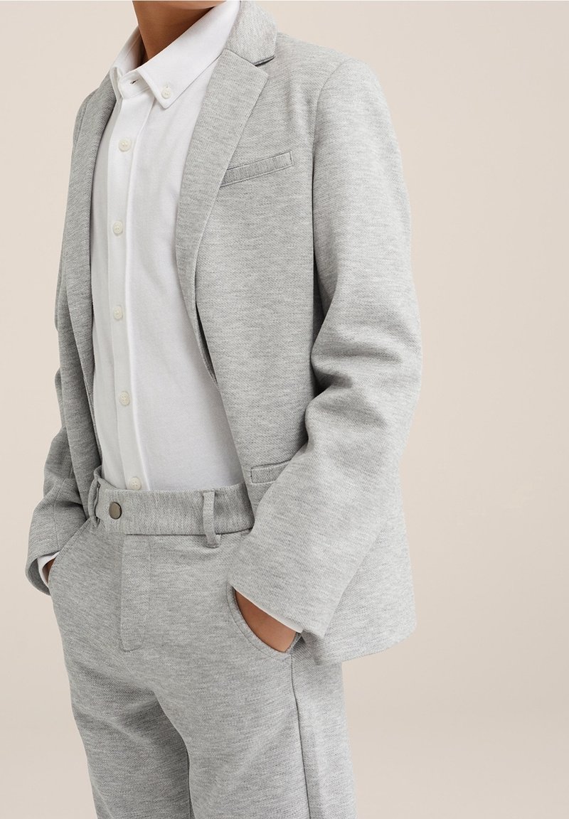 Person in einem hellgrauen Sakkowestens und einer hellgrauen Hose über einem weißen Hemd mit Knöpfen, die mit den Händen in den Taschen vor einem neutralen Hintergrund steht.