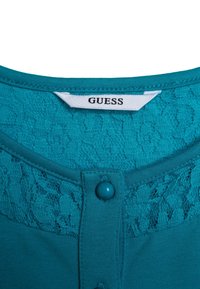 Błękitna bluzka z koronkowym wykończeniem przy kołnierzyku, okrągłym dekoltem i zapięciem na guziki. Wykonana z miękkiego, elastycznego materiału. Widoczna metka z marką.