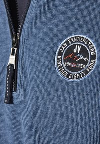 Blue zip-up sweater with a textured knit pattern, featuring a round black and white embroidered logo patch on the left chest.