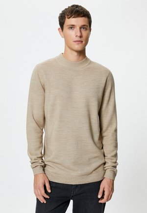 Suéter de punto beige con cuello redondo, puños acanalados y una textura sutil. El suéter tiene un corte relajado y una tela suave.