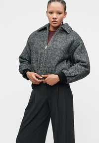 Veste à motif pied-de-poule, noire et grise, avec fermeture éclair et large col, associée à un pantalon sombre taille haute. Tissu texturé.