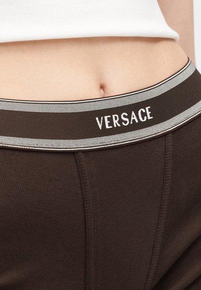 Κοντινό της μέσης ενός ατόμου που φορά σκούρα καφέ εσώρουχα με σήμα Versace και λευκό κολλητό τοπ που αποκαλύπτει την κοιλιά.