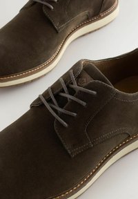 Acercamiento de zapatos de ante marrón oscuro con cordones, suelas de goma blanca y costuras detalladas sobre un fondo claro.