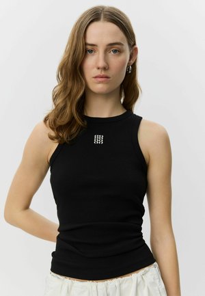 Junge Frau mit welligem hellbraunem Haar, trägt ein schwarzes ärmelloses Oberteil mit kleinem weißem Logo und weiße Hose vor einfarbigem Hintergrund.