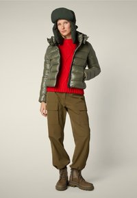 Giacca puffer verde oliva con zip frontale, maglione rosso a maglia, pantaloni cargo khaki e stivali marroni con lacci e suola spessa.