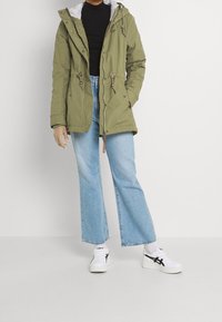 Olive green parka jacket with a gray fleece-lined hood, drawstring waist, and front pockets; paired with light blue wide-leg jeans and white sneakers.