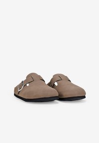 Mules marrones de cuero, sin talón, con punta redondeada, hebilla de metal ajustable y suela negra en contraste. Textura suave con cortes laterales.
