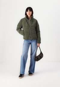Olijfgroene corduroy pufferjack met hoge kraag, ritssluiting en weerbestendig ontwerp, gecombineerd met lichtblauwe flare jeans en zwarte sneakers.