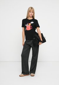 Sort t-shirt med et farverigt grafik af en tegneseriefigur i en festlig krus. Båret med grå videbukser og en sort taske.