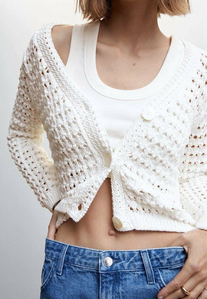 Femme portant un cardigan en crochet blanc par-dessus un débardeur blanc, associé à un jean bleu taille haute, mains posées sur les hanches.