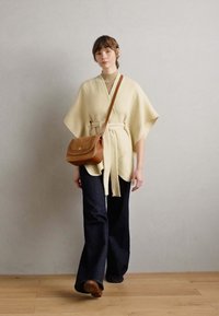 Beige poncho med brede ærmer, der er bælte om livet, sammen med mørke flared jeans og en brun crossbody taske. Glat, tekstureret stof.