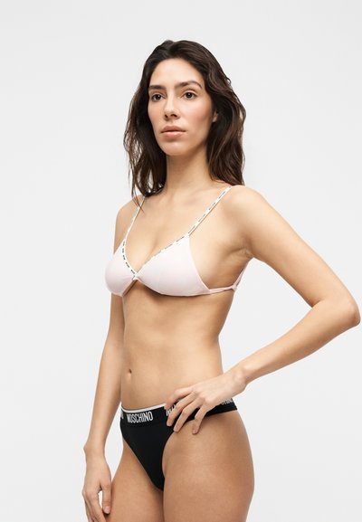 Γυναίκα με μακριά σκούρα μαλλιά που φοράει ροζ τριγωνικό σουτιέν και μαύρο σλιπ με λευκή κορδέλα "Moschino", στέκεται σε απλό ανοιχτό φόντο.