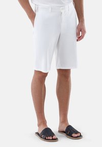 Weiße, knielange Shorts mit flachem Vorderteil, seitlichen Taschen und einem Knopfverschluss, kombiniert mit schwarzen Slide-Sandalen mit geflochtenen Riemen.