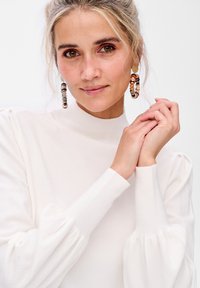 Biały sweter z wysokim kołnierzem i bufiastymi długimi rękawami; modelka nosi kolorowe kolczyki z koralików i ma jasne, falowane włosy, z dłońmi złożonymi na klatce piersiowej.