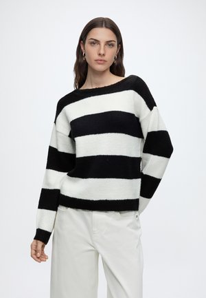 Femme aux longs cheveux foncés portant des boucles d’oreilles créoles dorées, un pull rayé noir et blanc, et un pantalon blanc sur un fond blanc uni.