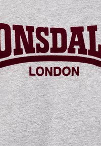 Pelēks sporta krekls ar mīkstu tekstūru, uz kura ir izteiksmīgs bordo krāsas teksts "LONSDALE" un zem tā, izliekumā, "LONDON".