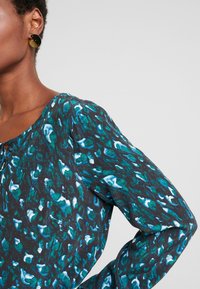 Czarny top z długim rękawem z abstrakcyjnym wzorem w odcieniach turkusu i bieli. Okrągły dekolt z wiązaniem z przodu, strukturalne wykończenie.