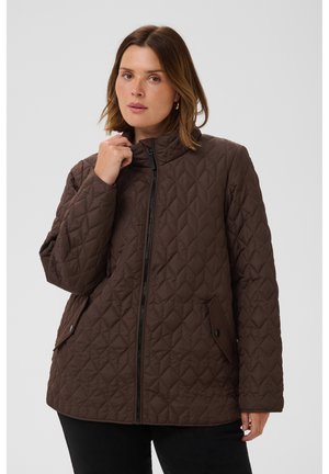 Femme aux cheveux mi-longs portant une veste matelassée marron avec une fermeture éclair devant et des poches à rabats, tenant le col d'une main.