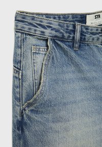 Jeans de mezclilla de color azul claro con acabado desgastado, con corte recto, dos bolsillos delanteros y una etiqueta cosida en la cinturilla. Detalle de tela texturizada visible.