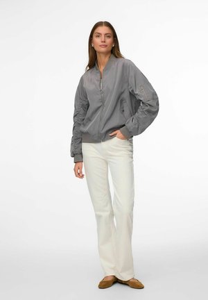 Femme portant une veste bomber grise zippée, un pantalon blanc droit et des ballerines marron, debout avec une main dans la poche sur fond blanc.