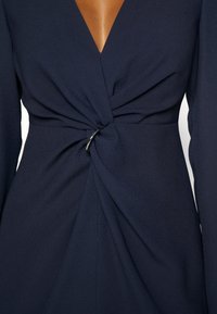 Robe bleu marine à manches longues, avec un décolleté en V profond et un détail de tissu torsadé maintenu par un anneau argenté à la taille.